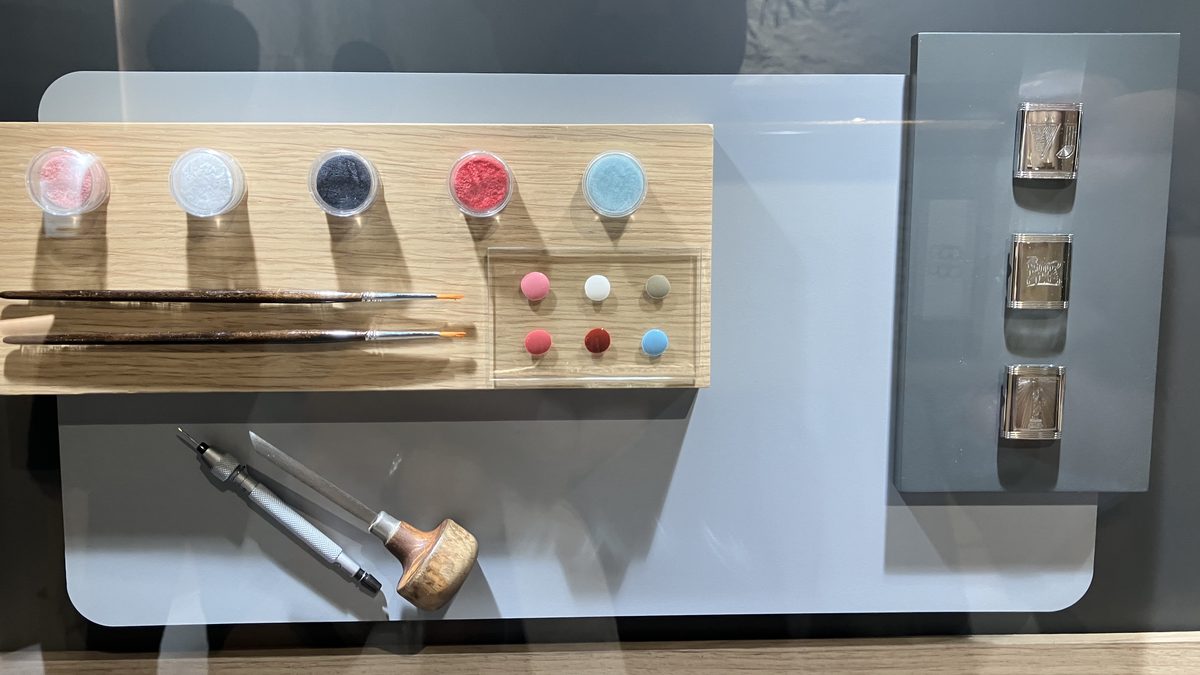 色別のエナメル顔料の容器、極細の筆、彫刻具を木製の台に並べた展示